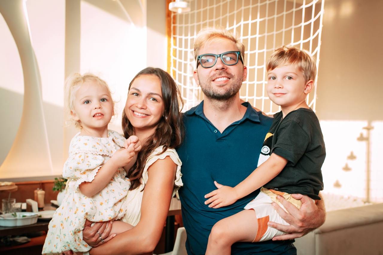 Stas y Masha con sus dos hijos en brazos, todos sonriendo en una cafetería